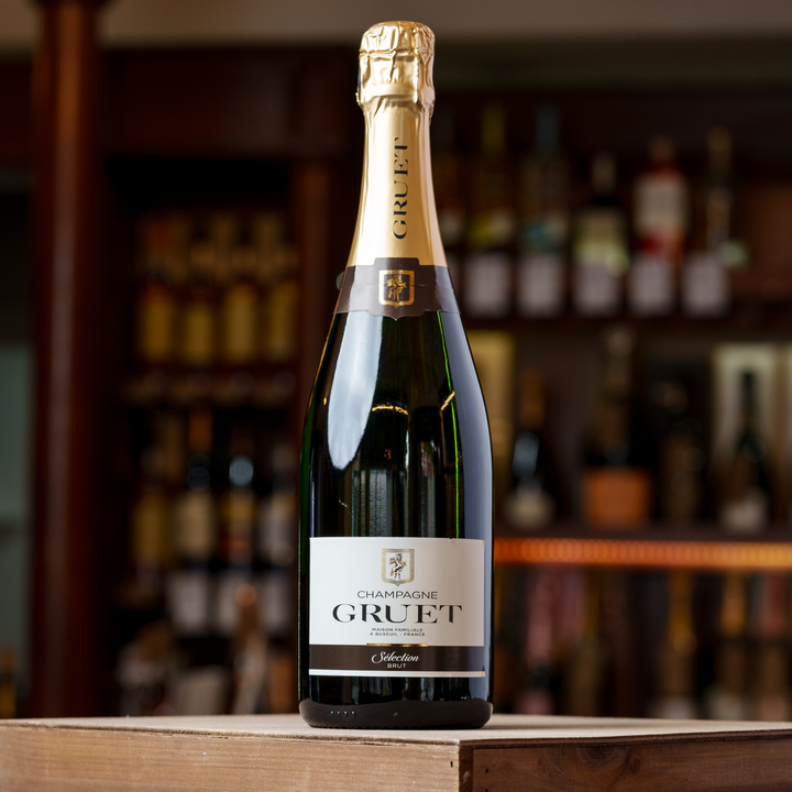 A bottle of Champagne Gruet Sélection Brut displayed on a wooden surface with a blurred background of premium liquor shelves, emphasizing its classic gold foil and elegant label design.