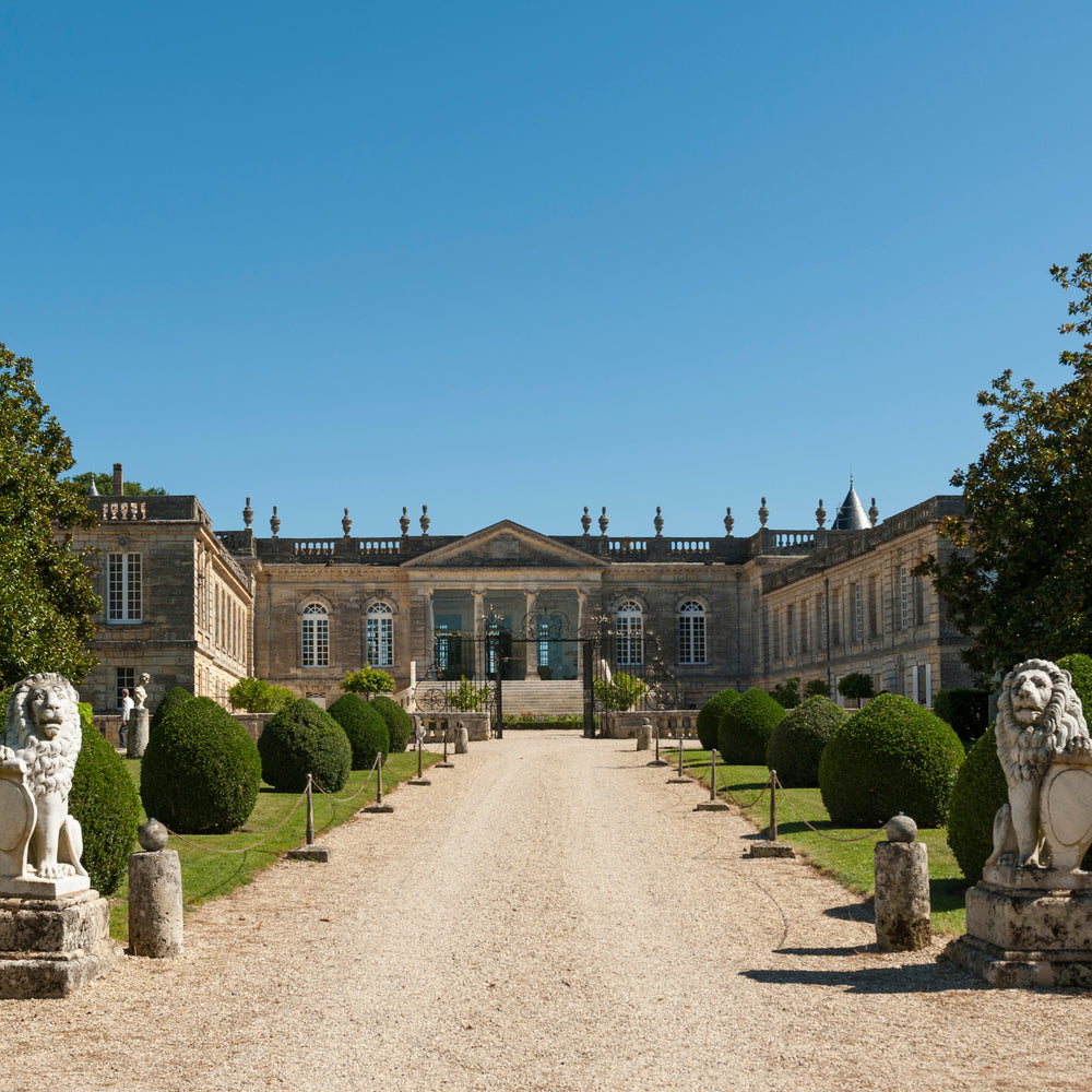 A grand view of Château St. Georges featuring its neoclassical architecture, flanked by meticulously maintained greenery and two regal lion statues guarding the entrance pathway under a clear blue sky.