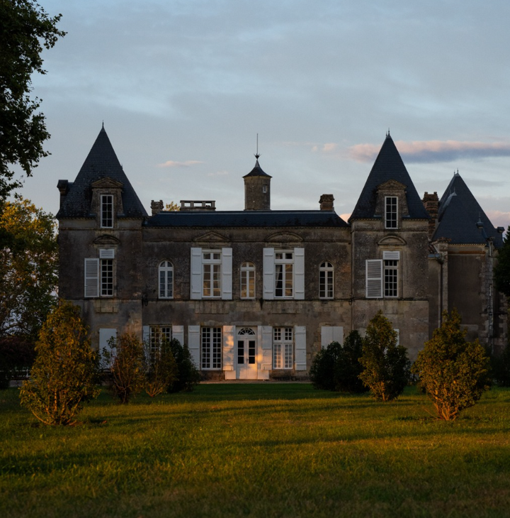 The historic façade of Château d'Issan at dusk, featuring towers and white shutters with the last sunlight illuminating the building.