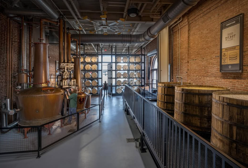 A view inside Michter's Distillery showcasing copper stills, wooden fermentation barrels, and rows of stacked barrels against a rustic brick wall.