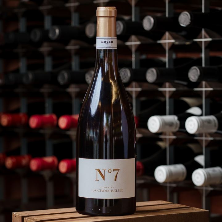 A premium bottle of Domaine La Croix Belle N°7 wine displayed on a wooden crate, set against a backdrop of neatly arranged wine bottles in a sophisticated wine cellar.