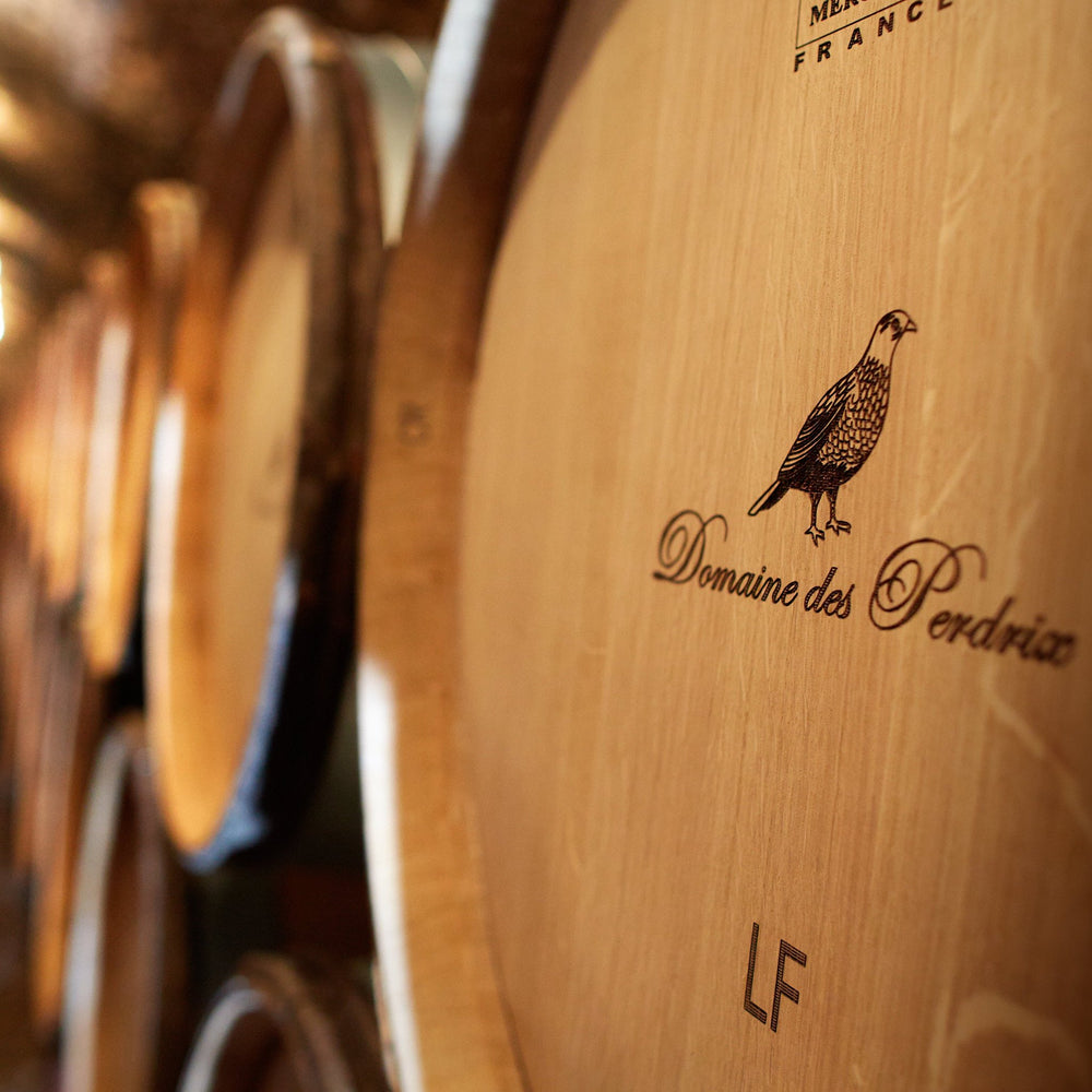 Close-up of a wooden wine barrel marked "Domaine des Perdrix" with an engraved partridge illustration, surrounded by rows of barrels in a dimly lit cellar.