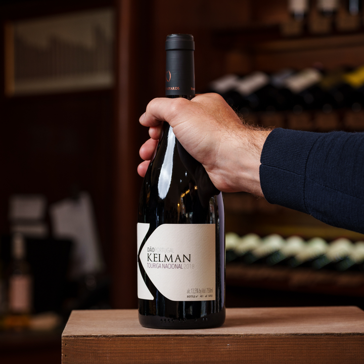 A hand holding a bottle of Kelman Touriga Nacional 2018 wine, showcased in a rustic setting with a blurred backdrop of a wine cellar, emphasizing its elegant and minimalist label design.