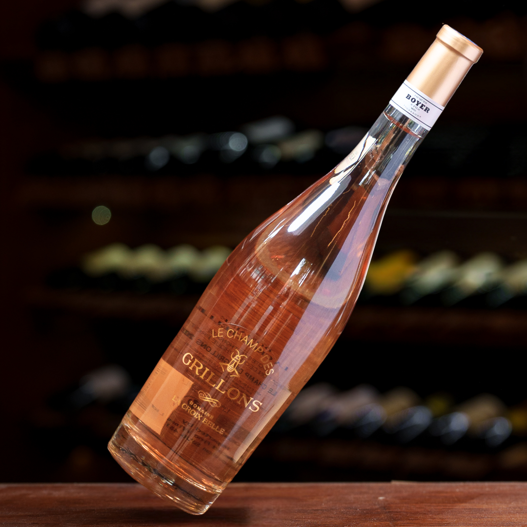 A sleek bottle of Le Champs des Grillons Rosé wine, elegantly tilted on a wooden surface with blurred wine racks in the background, emphasizing its premium quality and refined presentation.