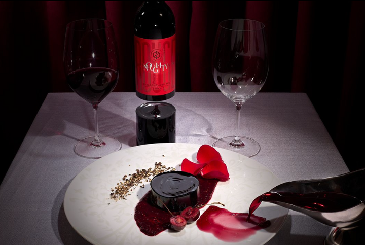 A bottle of Noughty Rouge Syrah dealcoholized red wine displayed alongside a gourmet plate, two wine glasses, and a poured glass of wine.