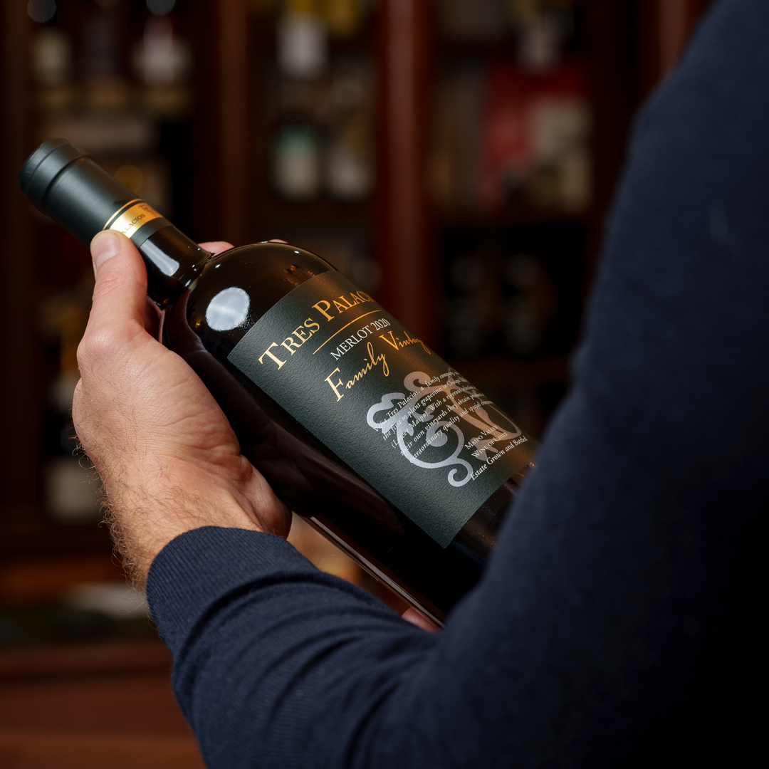 A person holding a bottle of Tres Palacios Merlot 2018, showcasing its elegant black and gold label design with a blurred wine display cabinet in the background, emphasizing its premium presentation.