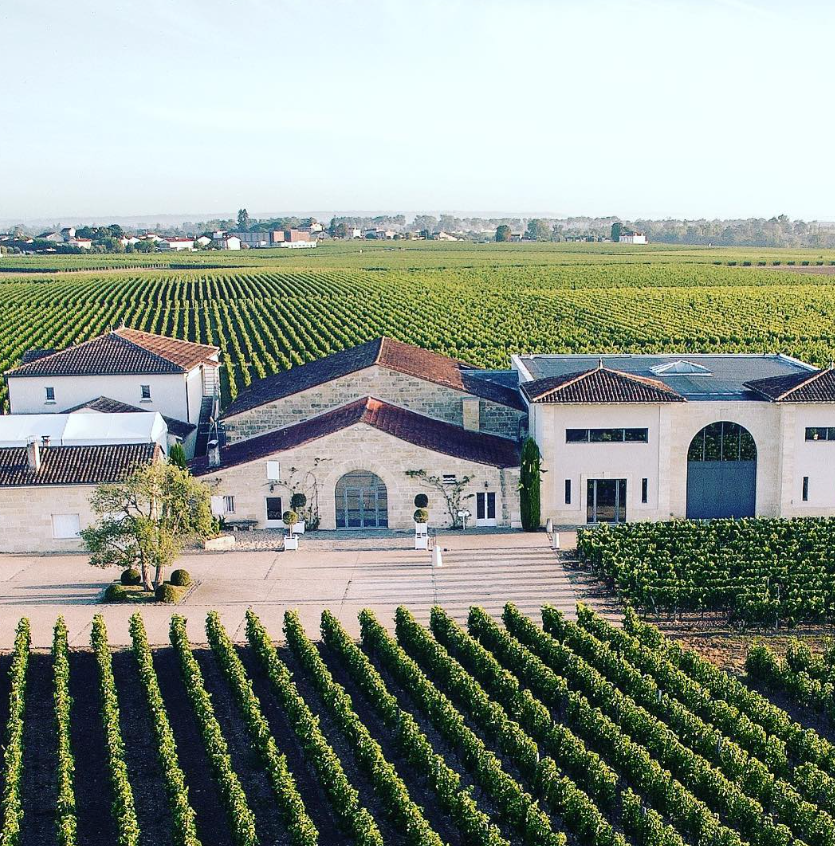 Aerial view of a La Conseillante winery surrounded by rows of lush green vineyards under a clear sky, showcasing the expansive vineyard estate.