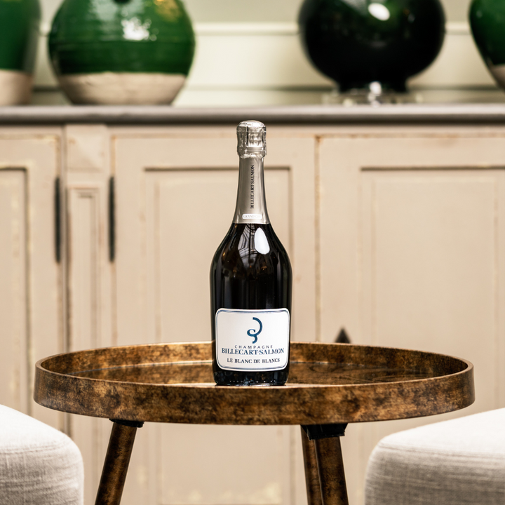 A bottle of Billecart-Salmon Le Blanc de Blancs Champagne elegantly displayed on a round, rustic table with a chic interior background featuring ceramic vases and a neutral cabinet.