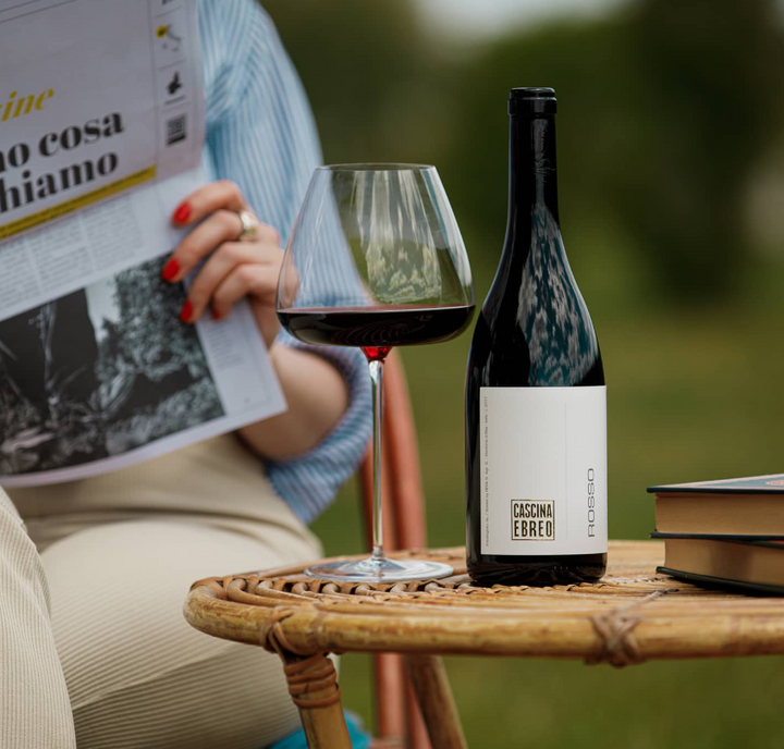 A Cascina Ebreo Rosso wine bottle on a wicker table, accompanied by a wine glass and books, in an outdoor setting.