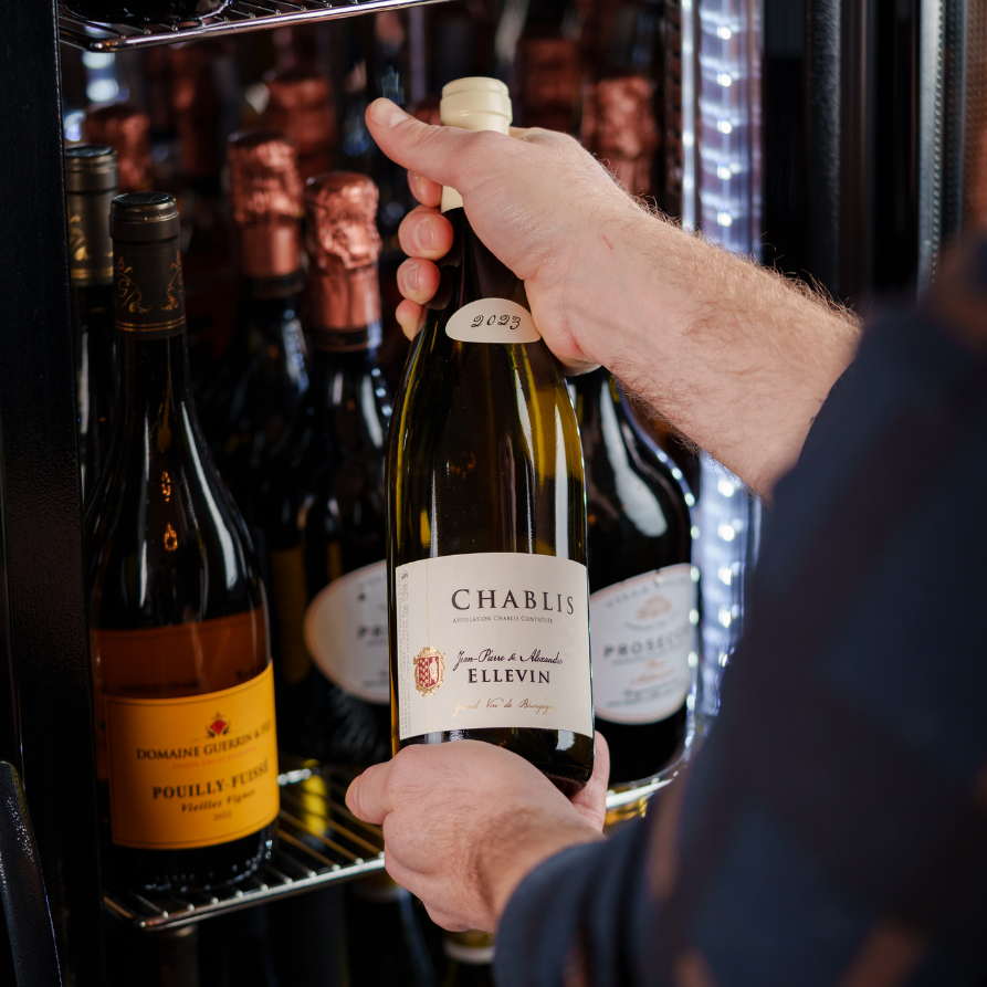 Hand selecting a bottle of Chablis Ellevin wine from a refrigerated shelf featuring premium wines, including Pouilly Fuissé and Prosecco bottles in the background.