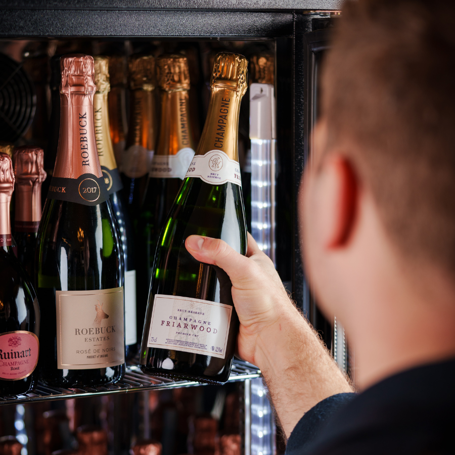 A person selecting a Friarwood champagne bottle from a wine fridge filled with various sparkling wine and champagne options, including Roebuck Estates and Ruinart labels.