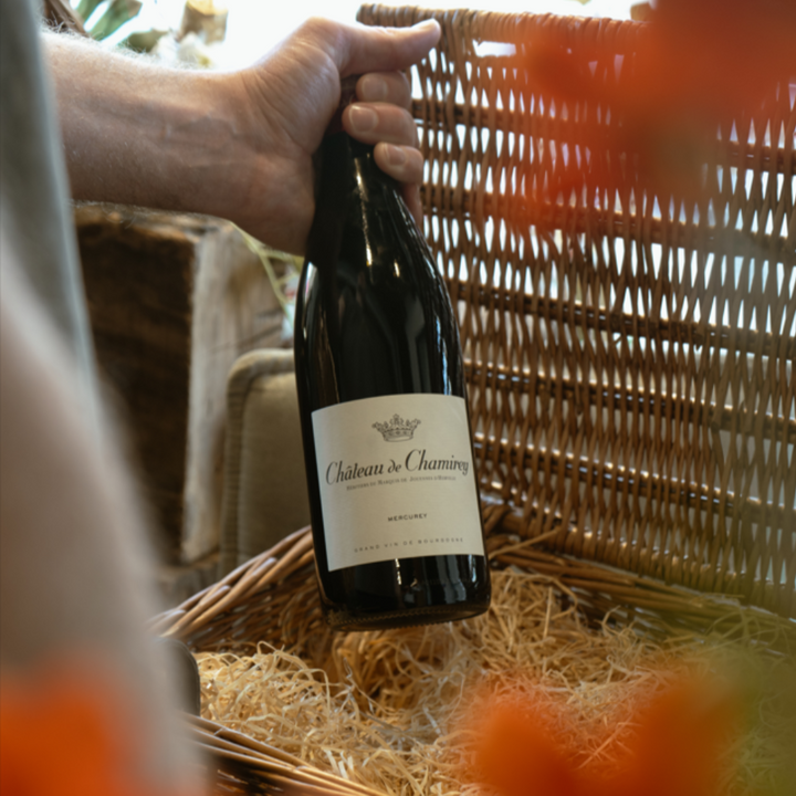 A hand holding a bottle of Château de Chamirey Mercurey wine, taking it out of a wicker basket.