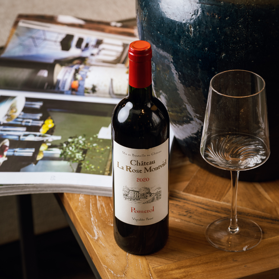 Château La Rose Montviel Pomerol 2020 wine bottle displayed on a wooden table next to a wine glass and an open magazine, showcasing a sophisticated tasting atmosphere.