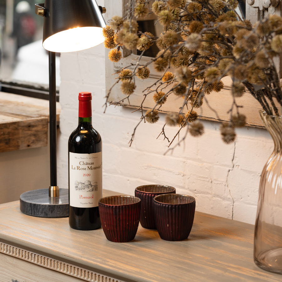 A bottle of Château La Rose Montviel Pomerol 2020 placed on a rustic wooden table next to a lamp, decorative cups, and dried flowers, creating a cozy, elegant setting.