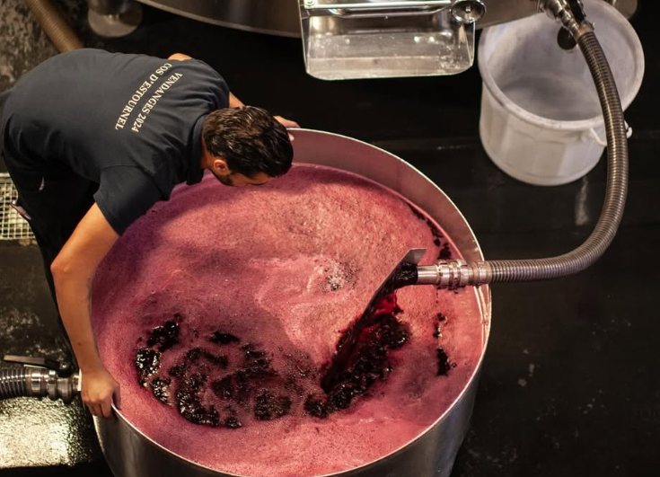 A winemaker overseeing the fermentation process at Cos d'Estournel, with a large vat of vibrant red wine.