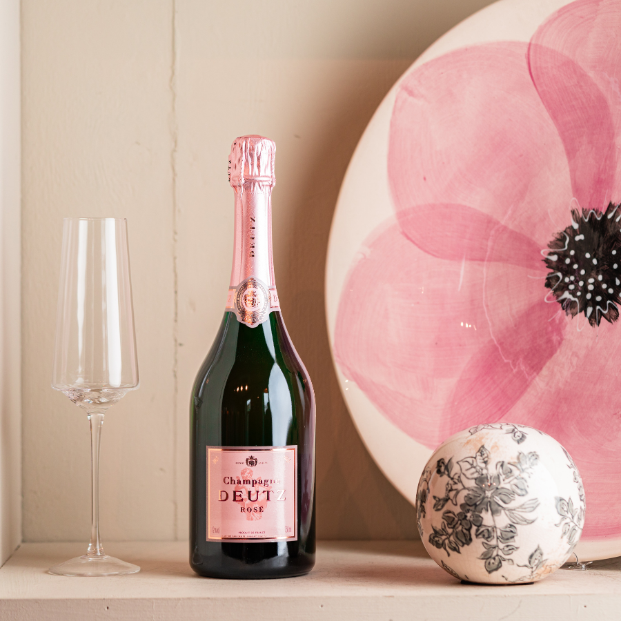 A bottle of Deutz Rosé Champagne displayed on a decorative shelf with a flute glass, a floral ceramic plate, and a textured ornament, highlighting an elegant and refined setting.