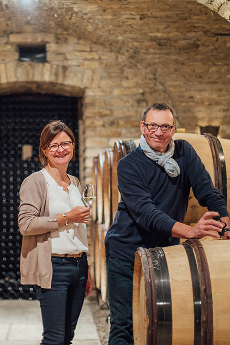 Aline and Vincent Bouzereau standing in their wine cellar with barrels, holding glasses of wine.