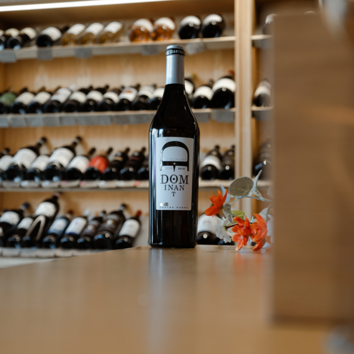 A Dominant white wine bottle placed on a wooden counter in a wine shop with shelves of wine bottles in the background.