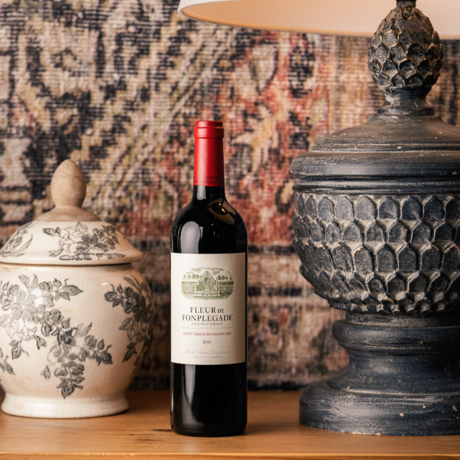 Fleur de Fonplégade 2018 Saint-Émilion Grand Cru wine bottle placed on a wooden shelf next to a ceramic jar and a rustic lamp, against a tapestry backdrop, emphasizing a luxurious and artisan aesthetic.