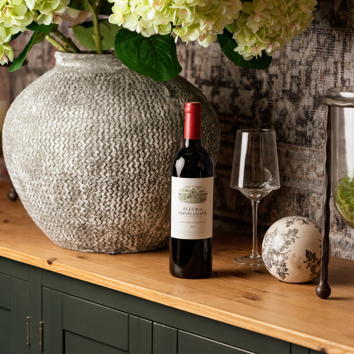 A bottle of Fleur de Fonplégade 2018 Saint-Émilion Grand Cru wine displayed on a wooden counter with a large vase of hydrangeas, a wine glass, and decorative elements, creating an elegant setting.