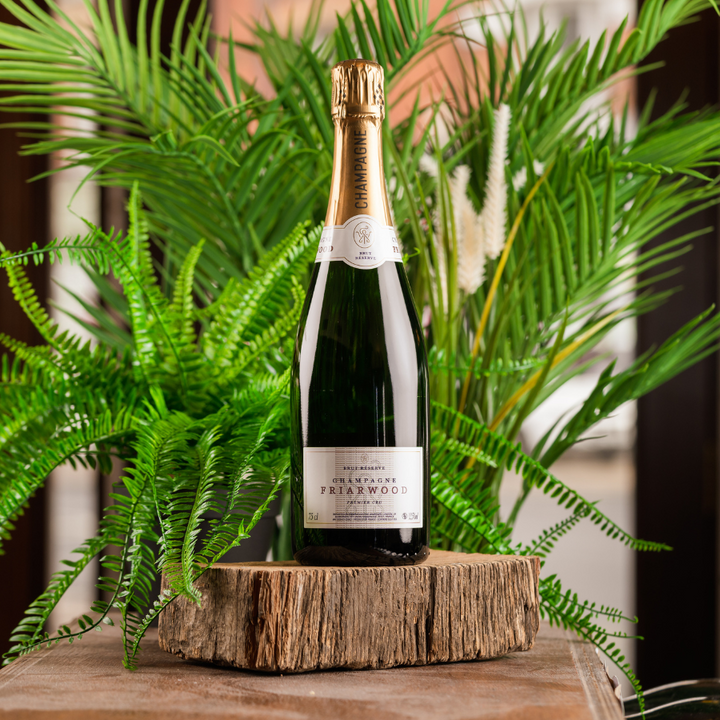 A bottle of Friarwood Champagne on a wooden slice surrounded by greenery.