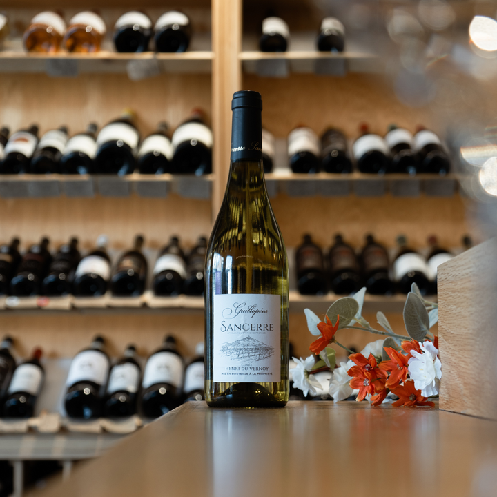 A bottle of Guillopees Sancerre displayed on a polished wooden counter, adorned with fresh orange and white flowers, with a backdrop of a wine cellar featuring rows of bottles.