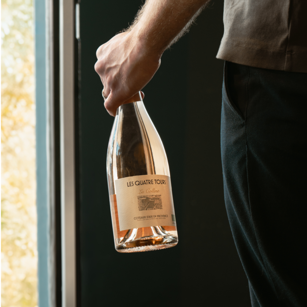 A hand holding a bottle of Les Quatre Tours La Colline Rosé wine near a window.