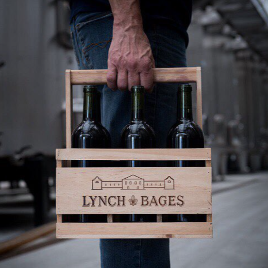 A wooden crate labeled "Lynch Bages" containing three wine bottles, held by a hand in a winery setting.