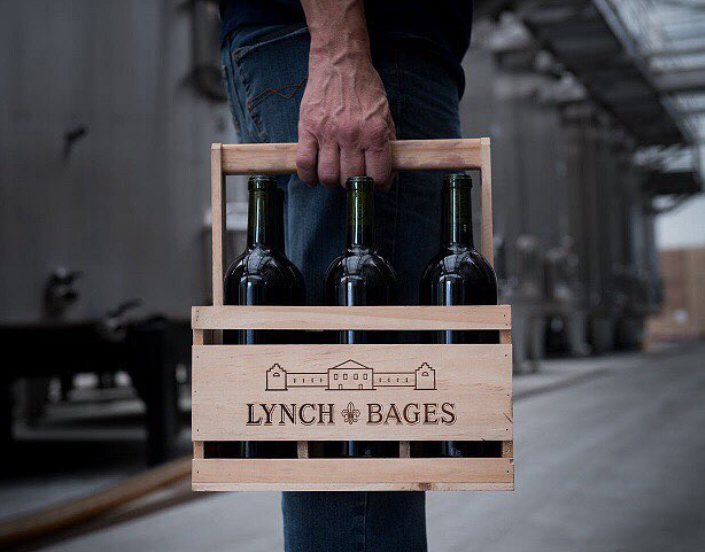 A wooden crate labeled "Lynch Bages" containing three wine bottles, held by a hand in a winery setting.