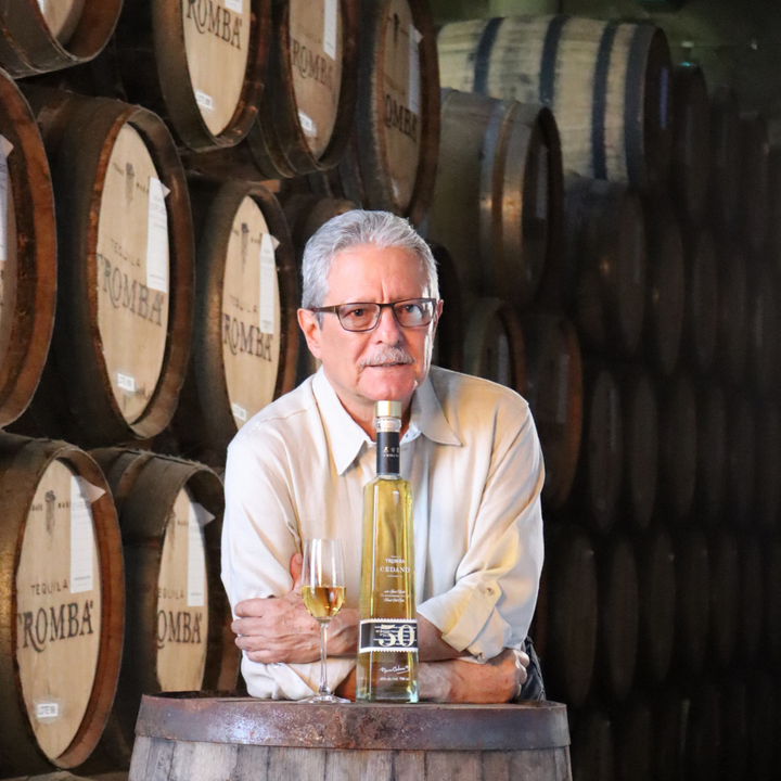 Master distiller Marco Cedano standing in front of Tromba Tequila barrels, showcasing a bottle of Tromba Cedano Reposado.