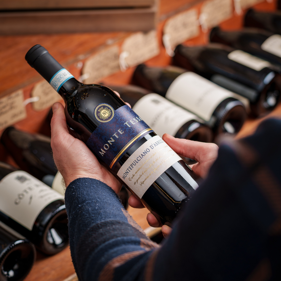 Hands holding a bottle of Monte Tessa Montepulciano d'Abruzzo wine in a wine store, with a background of neatly arranged wine bottles and handwritten shelf labels.