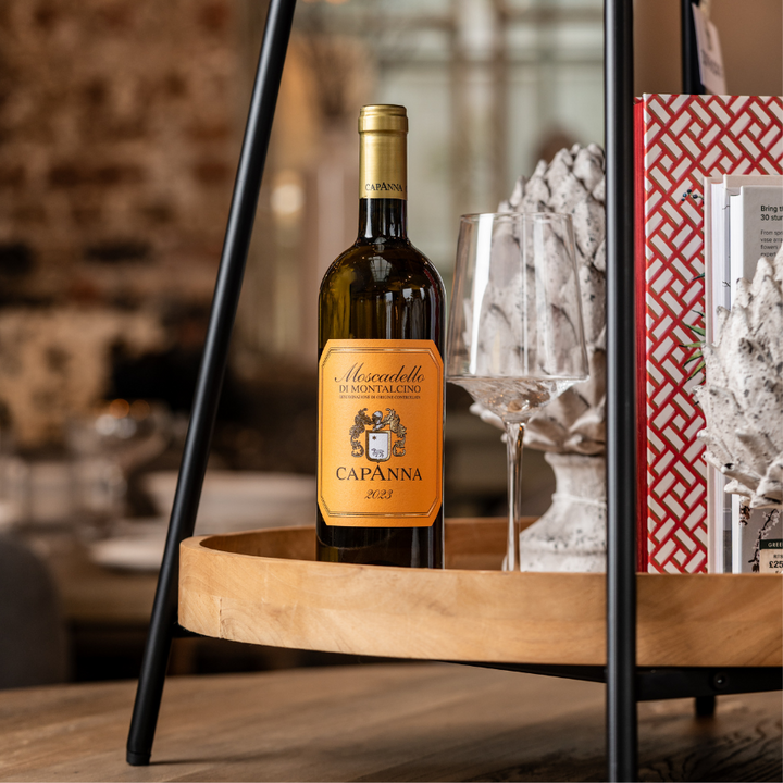 A bottle of Moscadello di Montalcino Capanna 2023 displayed on a wooden tray with wine glasses and decorative elements, creating an inviting and refined wine setting.