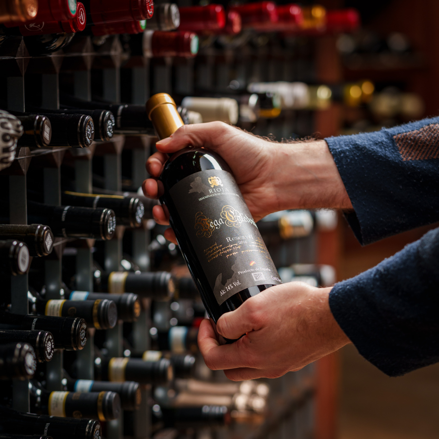 Close-up of hands holding a Rioja Reserva wine bottle in a wine store, highlighting the label and luxury ambiance amidst a background of wine racks.