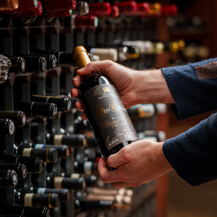 Close-up of hands holding a Rioja Reserva wine bottle in a wine store, highlighting the label and luxury ambiance amidst a background of wine racks.