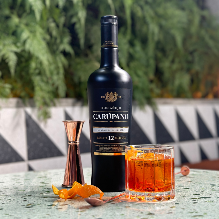 Ron Carúpano Reserva 12 Exclusiva bottle alongside a glass of rum cocktail garnished with an orange twist on a textured table.