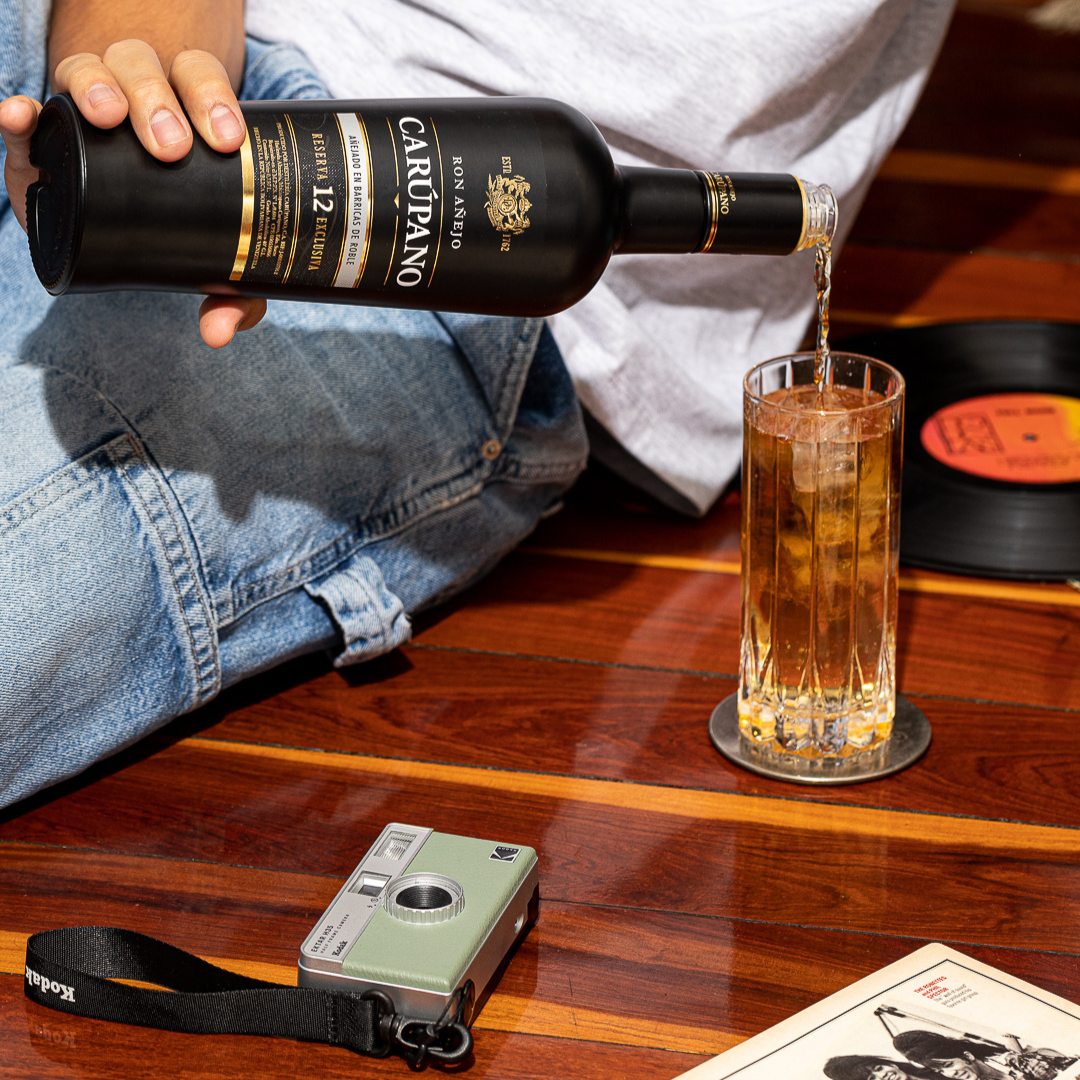 Person pouring Ron Carúpano Reserva 12 Exclusiva rum into a tall glass with ice, placed on a wooden surface alongside a vintage camera and vinyl record.