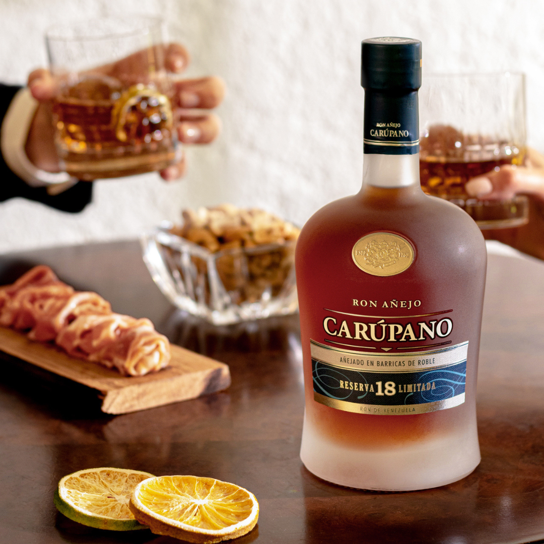 Ron Carúpano Reserva 18 Limitada bottle displayed on a wooden table, surrounded by dried citrus slices, snacks, and people enjoying glasses of rum.