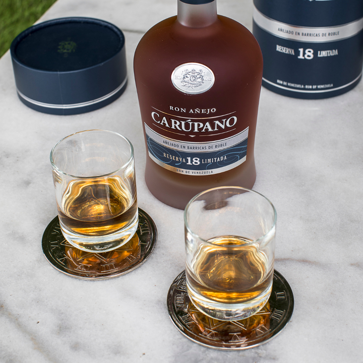 Ron Carúpano Reserva 18 Limitada bottle alongside two glasses of rum on decorative coasters, set on a marble surface.