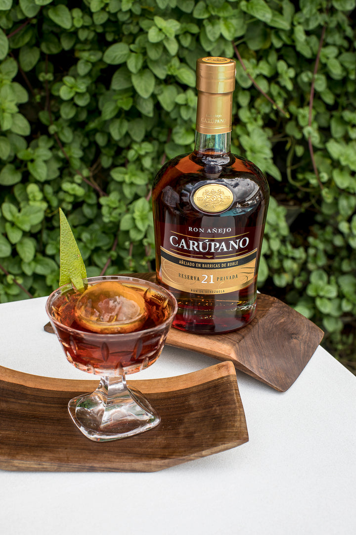 Ron Carúpano Reserva 21 Privada bottle alongside a glass of rum with ice and lime garnish, displayed on wooden trays against a green foliage background.