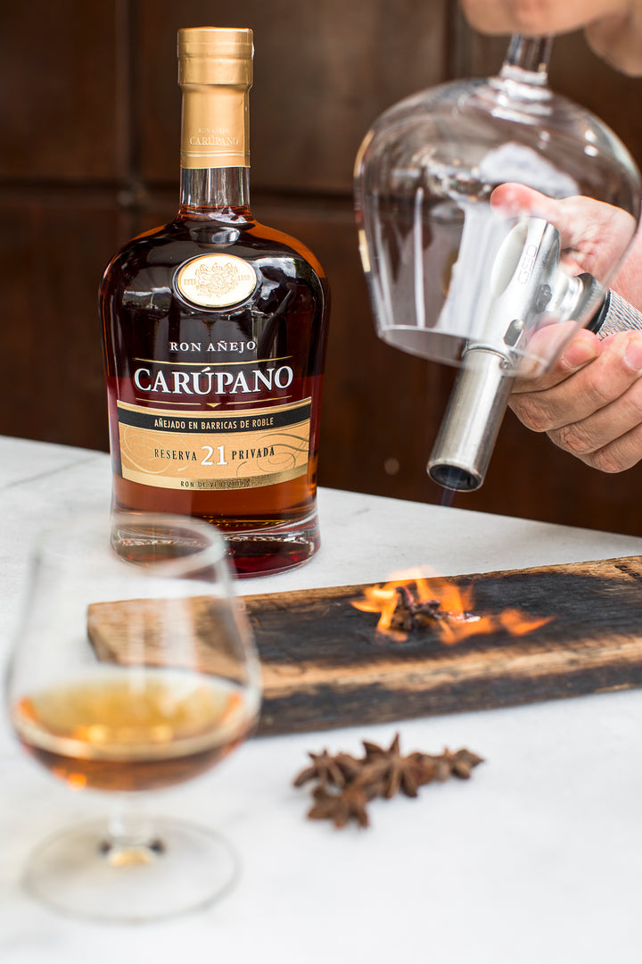 Ron Carúpano Reserva 21 Privada bottle with a glass of rum, flaming wood, and star anise, featuring a bartender using a smoking gun for presentation.