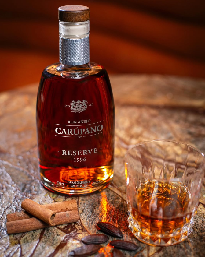 A bottle of Ron Carúpano Reserve 1996 rum on a textured surface, accompanied by a glass of rum, cinnamon sticks, and cocoa beans.