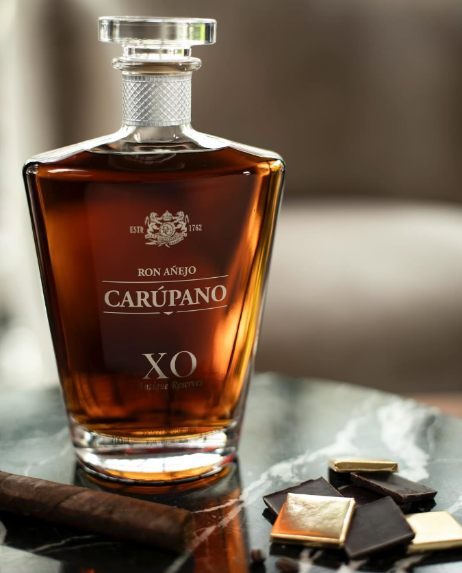 A bottle of Ron Carúpano XO Unique Reserves rum on a marble surface, accompanied by dark chocolate, gold-wrapped pieces, and a cigar.