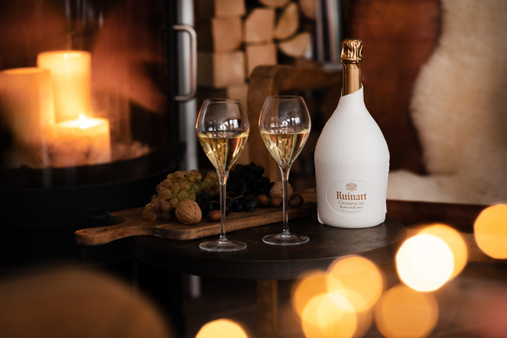 Ruinart Blanc de Blancs Champagne elegantly displayed by a cozy fireplace with grapes, walnuts, and sparkling flutes.