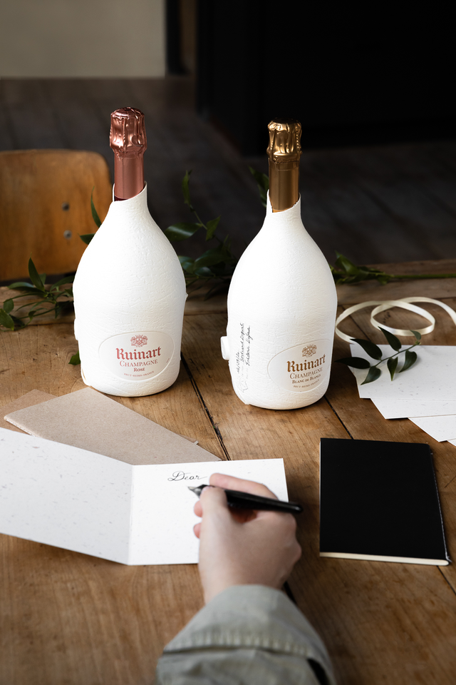 Two Ruinart Champagne bottles, elegantly wrapped in white sustainable packaging, sitting on a wooden table with a handwritten card.