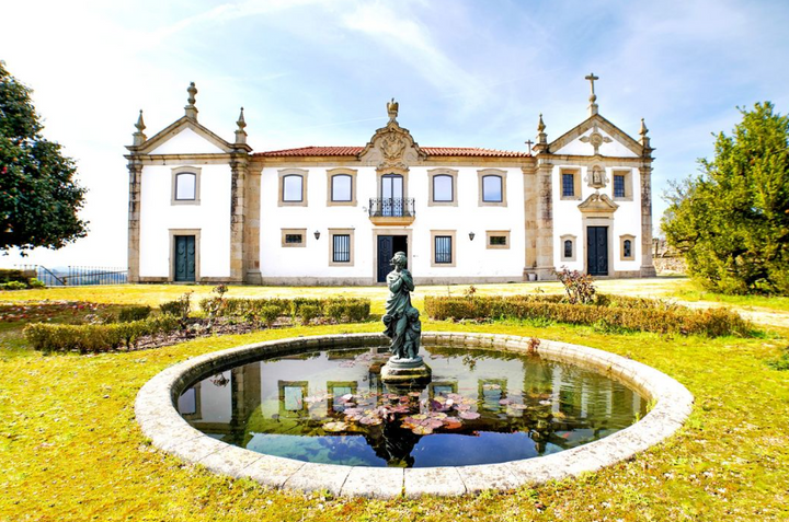 The historic mansion of Solar das Bouças estate, featuring a fountain with a statue, a garden, and a bright blue sky.