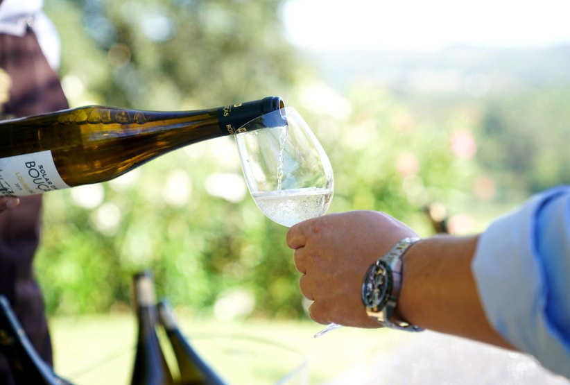 A close-up of a wine bottle pouring white wine into a glass outdoors, with a scenic green background.