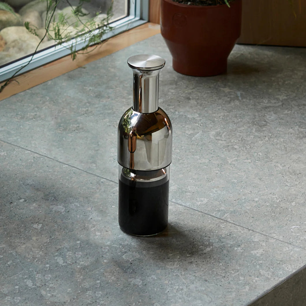 Sleek stainless steel eto wine decanter with a modern mirrored finish, elegantly displayed on a stone countertop near a window with potted plants.