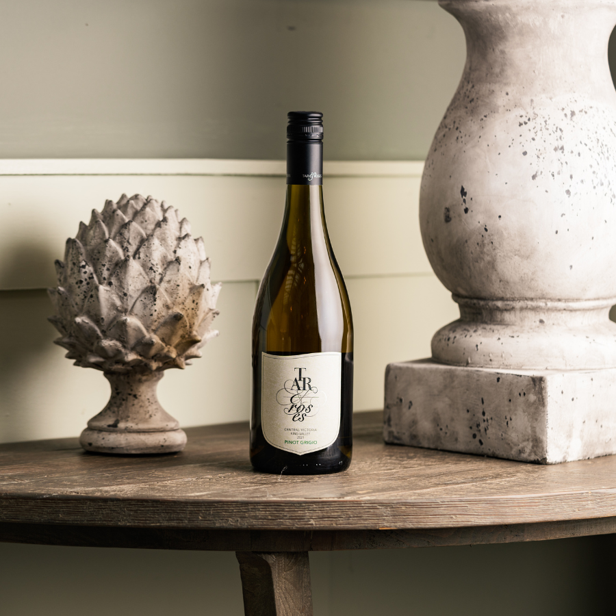 A bottle of Tar and Roses Pinot Grigio displayed on a wooden table with stone ornaments in a rustic setting, creating an elegant and earthy wine presentation.