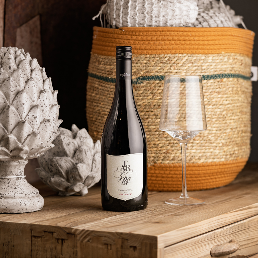 A bottle of Tar and Roses Sangiovese wine displayed on a wooden table with a wine glass, stone decorative ornaments, and a woven basket in the background, creating a rustic and cozy atmosphere.