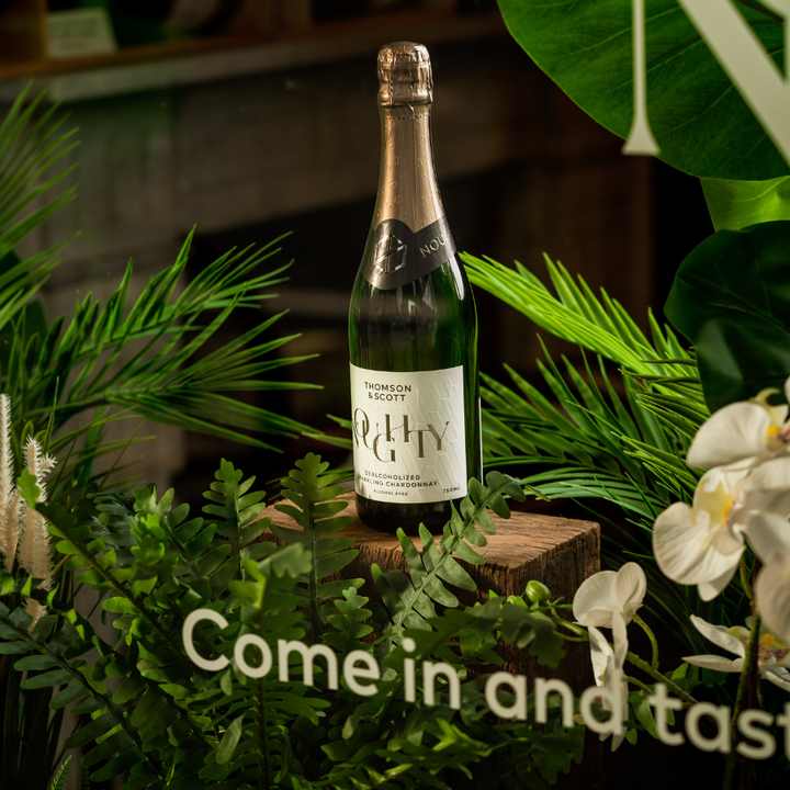 A bottle of Thomson & Scott Noughty Organic Sparkling Chardonnay on a wooden crate with a "Come in and taste" sign.